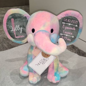 May include: A tie-dye plush elephant with gray ears. The ears have embroidered details, one ear says "Lilly" and the other says "08.10.2021 8 pounds 1 ounce 11:29 am". The elephant is sitting on a white surface with a tag that says "Nugs & Kisses".