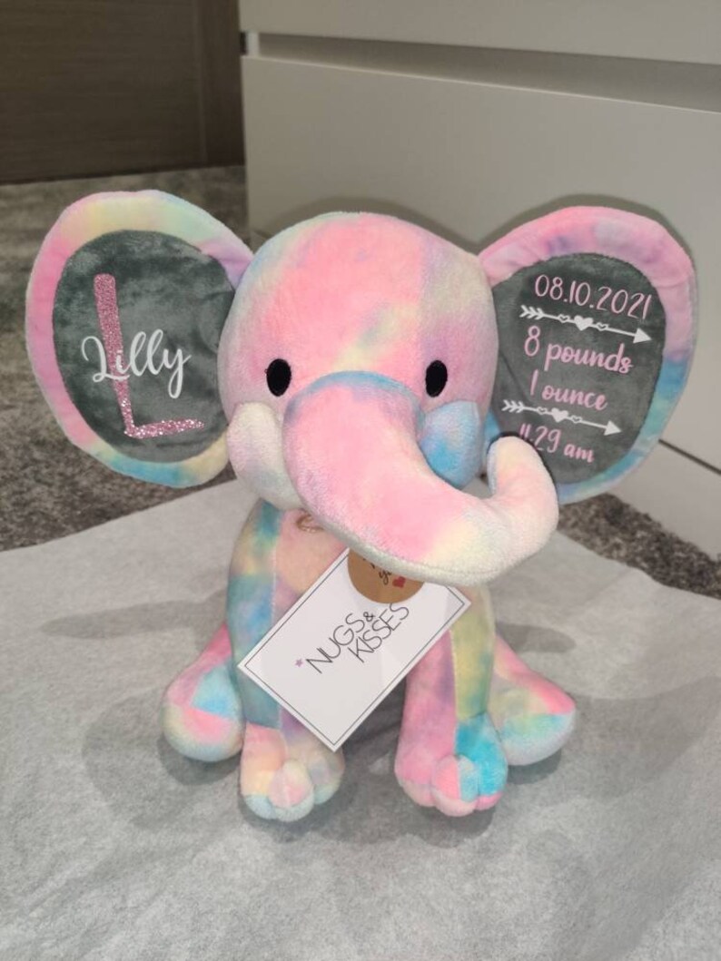 May include: A tie-dye plush elephant with gray ears. The ears have embroidered details, one ear says "Lilly" and the other says "08.10.2021 8 pounds 1 ounce 11:29 am". The elephant is sitting on a white surface with a tag that says "Nugs & Kisses".