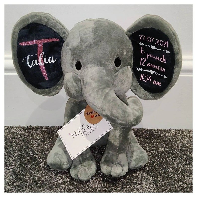 May include: Gray plush elephant with black ears. The ears have pink glitter writing. One ear says "Talia" and the other ear says "27.07.2021 6 pounds 12 ounces 11:54 am". A white tag with black writing says "NUGS & KISSES".