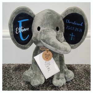Op de afbeelding: Een grijze pluche olifant met zwarte accenten op de oren. Op het linkeroor staat de naam "Ethan" in wit en blauw. Op het rechteroor staat "Christened 25.07.2021" met een kruis. Een label leest "Thank you!" en "Hugs & Kisses".