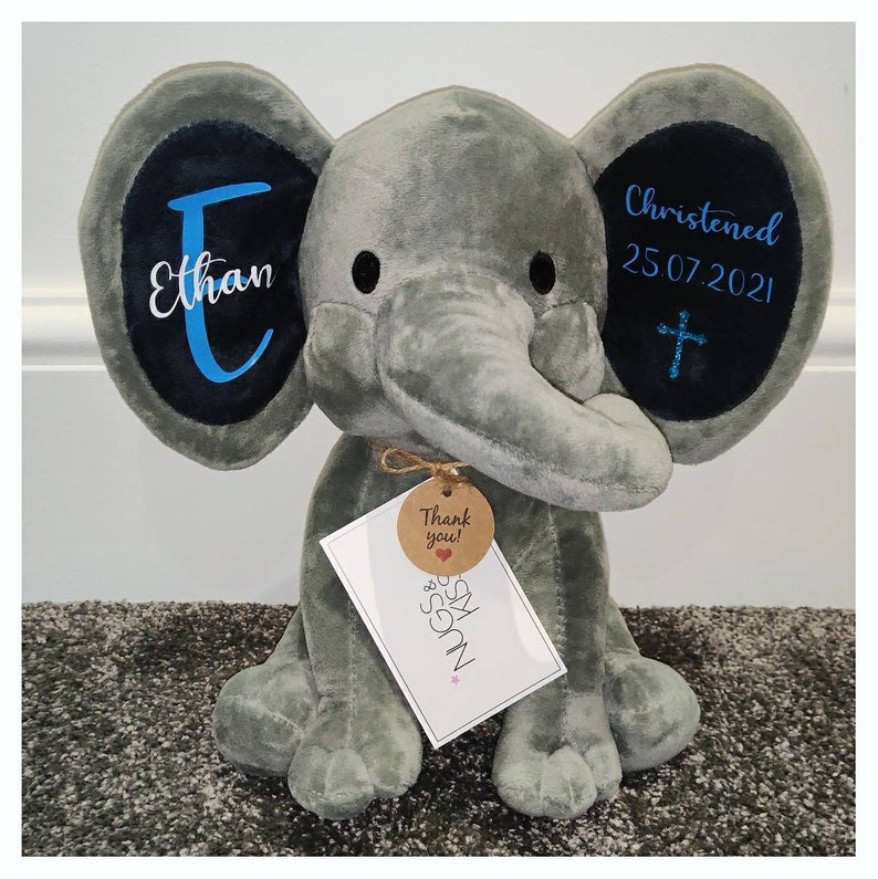 Op de afbeelding: Een grijze pluche olifant met zwarte accenten op de oren. Op het linkeroor staat de naam "Ethan" in wit en blauw. Op het rechteroor staat "Christened 25.07.2021" met een kruis. Een label leest "Thank you!" en "Hugs & Kisses".
