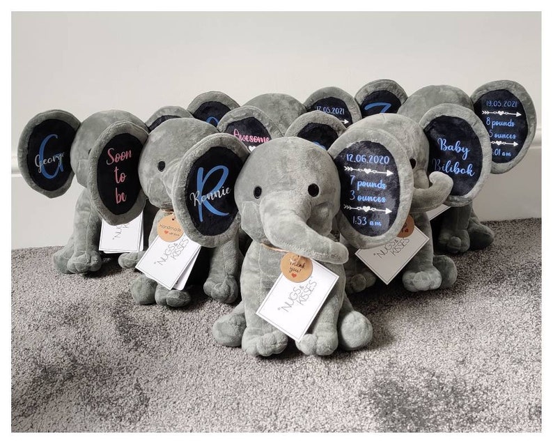 May include: A group of gray plush elephant toys with black ears. Each elephant has a personalized embroidered message on its ear. The messages include names, birth dates, weights, and times. The elephants are arranged in a row on a gray carpet.