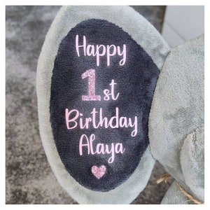 Op de afbeelding: Een zachte grijze olifanten oor met een donkergrijze binnenkant. De tekst "Happy 1st Birthday Alaya" staat in roze letters, met een glitterroze "1st" en een roze hart. Een leuk cadeau voor een eerste verjaardag.