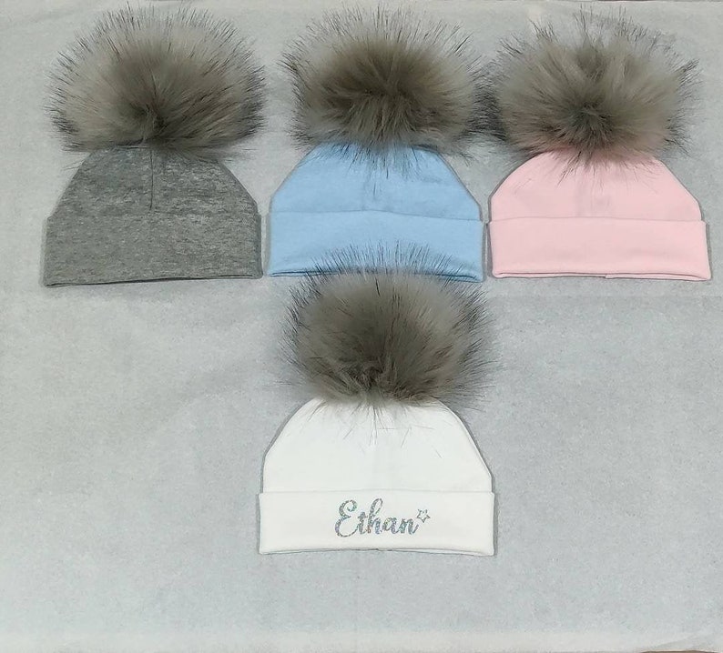 May include: Four knit hats with faux fur pom poms. Three hats are solid colors: gray, light blue, and pink. The fourth hat is white with the name "Ethan" embroidered in silver glitter.