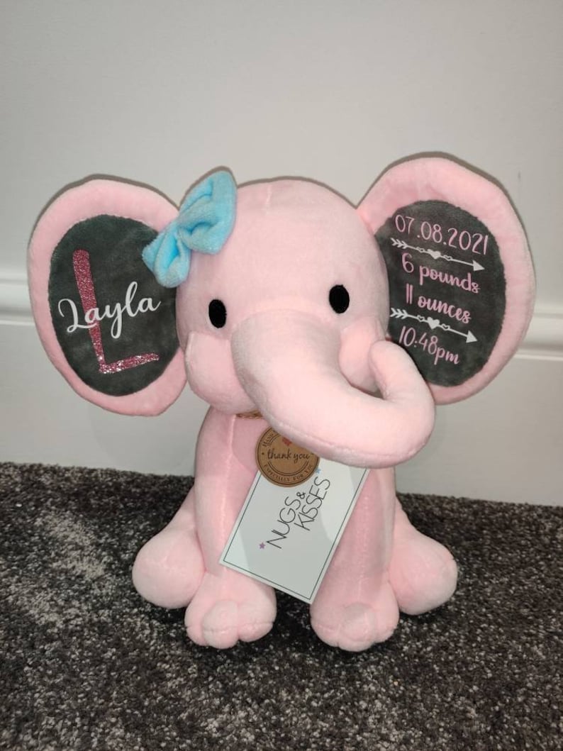 May include: Pink plush elephant stuffed animal with a gray ear tag that says "Layla" and another gray ear tag that says "07.08.2021 6 pounds 11 ounces 10:48pm". The elephant is holding a small brown tag that says "Thank you for the NUGS & KISSES".