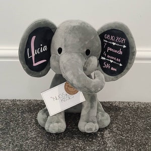 May include: Gray plush elephant stuffed animal with black ears. The ears have white writing on them. One ear says "Lucia" in pink lettering. The other ear says "08.10.2021 7 pounds 4 ounces 5:14 am". The elephant is sitting on a gray carpet. A white tag with the words "NUGS & KISSES" is attached to the elephant.