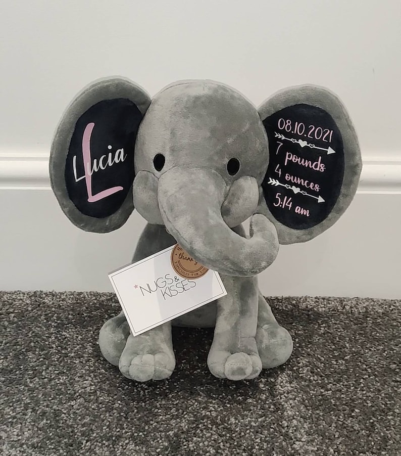 May include: Gray plush elephant stuffed animal with black ears. The ears have white writing on them. One ear says "Lucia" in pink lettering. The other ear says "08.10.2021 7 pounds 4 ounces 5:14 am". The elephant is sitting on a gray carpet. A white tag with the words "NUGS & KISSES" is attached to the elephant.