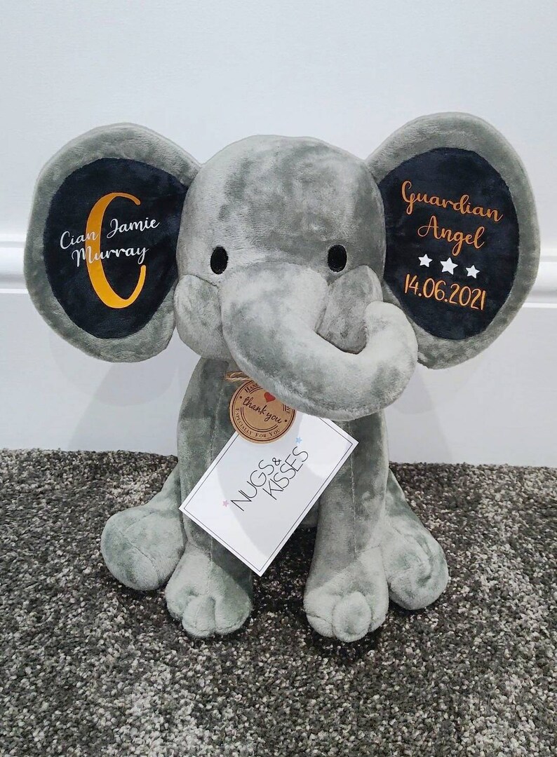 Op de afbeelding: Een grijze pluche olifant met geborduurde details op de oren. Op &eacute;&eacute;n oor staat "Cian Jamie Murray" en op het andere "Guardian Angel" met een datum. Een label leest "Nugs & Kisses".