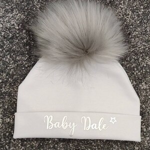 May include: White baby hat with a fluffy white pom pom and the text "Baby Dale" embroidered in silver on the front.