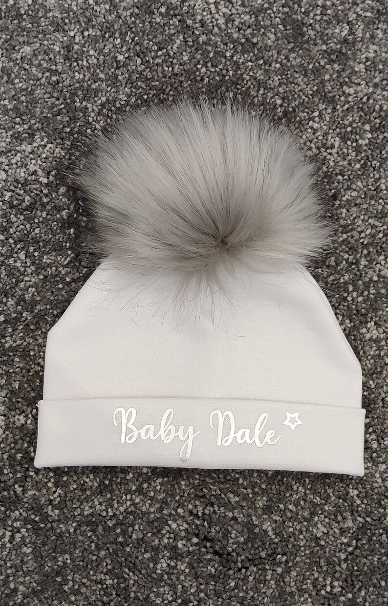 May include: White baby hat with a fluffy white pom pom and the text "Baby Dale" embroidered in silver on the front.