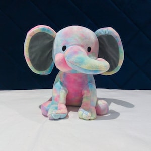 May include: A plush toy elephant with a tie-dye pattern in shades of pink, blue, and yellow. The elephant has large gray ears and a small trunk.