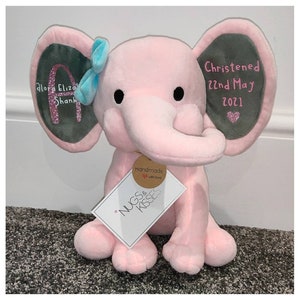 Op de afbeelding: Een roze pluche olifant met grijze ooraccenten en een lichtblauwe strik. De oren zijn geborduurd met tekst en een glitterletter. Een label leest "Nugs & Kisses" en "Handmade with love."