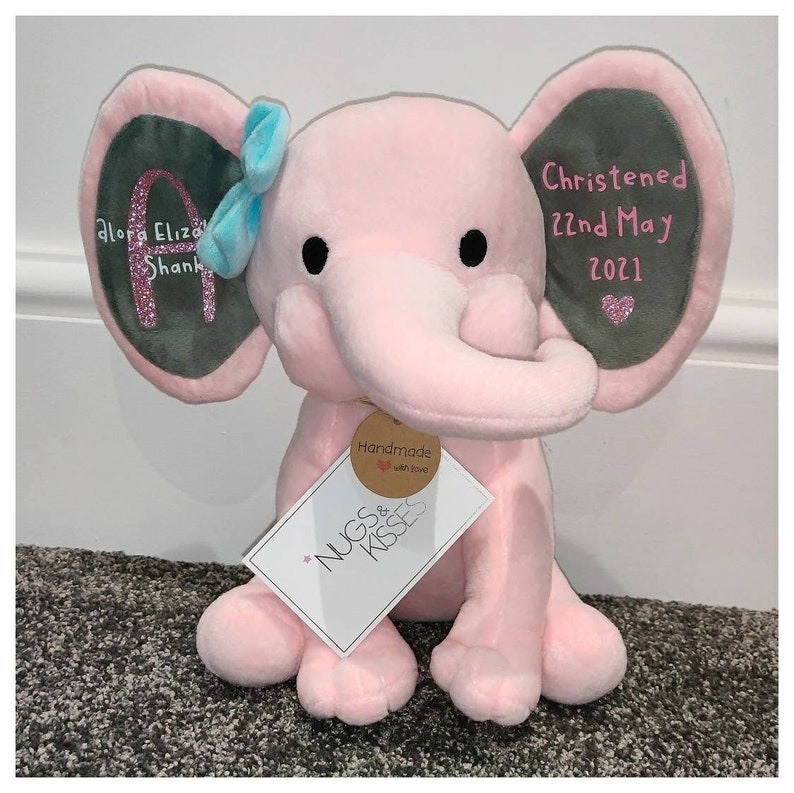 Op de afbeelding: Een roze pluche olifant met grijze ooraccenten en een lichtblauwe strik. De oren zijn geborduurd met tekst en een glitterletter. Een label leest "Nugs & Kisses" en "Handmade with love."