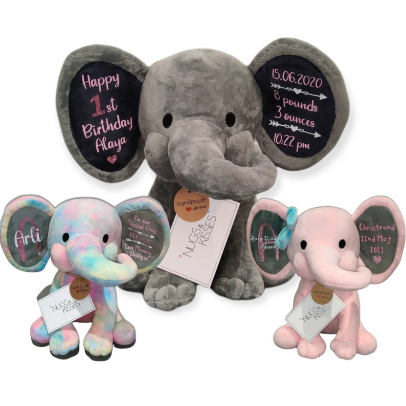 Op de afbeelding: Drie pluche olifanten in grijs, pastel regenboog en roze. De grijze olifant heeft geborduurde tekst op zijn oor met de tekst "Happy 1st Birthday Alaya". De andere olifanten hebben gepersonaliseerde tekst op hun oren en labels met "Nugs & Kisses".