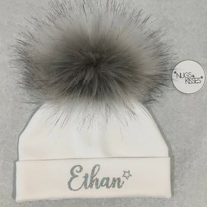 May include: White baby hat with a fluffy gray pom pom and the name "Ethan" embroidered in silver glitter on the front. The hat has a small tag that says "NUGS & KISSES".