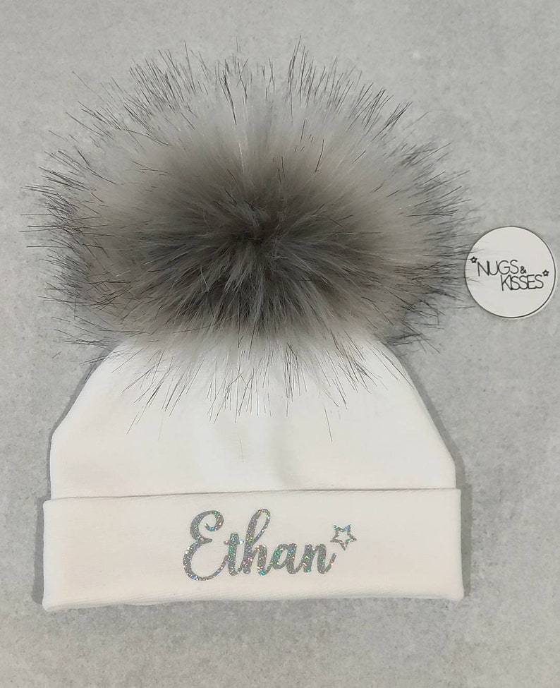 May include: White baby hat with a fluffy gray pom pom and the name "Ethan" embroidered in silver glitter on the front. The hat has a small tag that says "NUGS & KISSES".