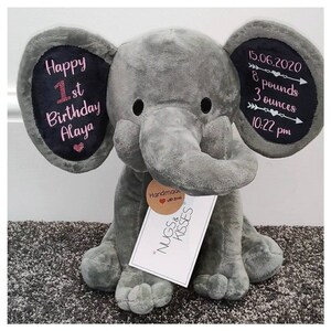 May include: Gray plush elephant stuffed animal with embroidered details on the ears. One ear says "Happy 1st Birthday Alaya" and the other ear says "15.06.2020 8 pounds 3 ounces 10:22 pm". A small brown tag with a heart and the text "Handmade with love" is attached to the elephant's leg. A white tag with pink stars and the text "NUGS & KISSES" is attached to the elephant's leg.