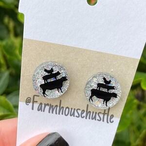 May include: A pair of silver glitter stud earrings with a black silhouette of a cow, pig, and chicken stacked on top of each other. The earrings are on a white card with the text "@farmhousehustle".