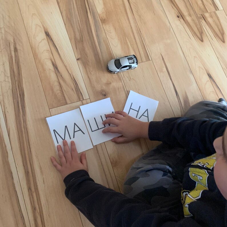 May include: A child is learning to spell the Russian word "машина" (car) by arranging letter tiles on a wooden floor. The tiles are white with black lettering. The child is holding the tile with the letter "Ш" in their hand. A small toy car is in the background.