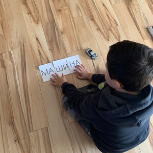 May include: A child sits on a wooden floor and spells out the word "МАШИНА" (Russian for "car") with letter tiles. A toy car is on the floor next to the tiles.