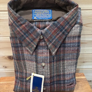 May include: A folded Pendleton button-down shirt in a plaid pattern of brown, red, and blue. The shirt has a collar and buttons down the front. A tag reads "Genuine Pendleton 100% Virgin Wool" and "Made in USA."