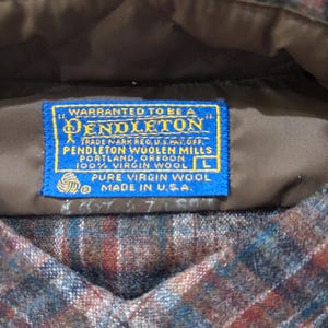May include: Close-up of a Pendleton wool shirt label. The blue label reads "Warranted to be a Pendleton" and "100% Virgin Wool." The shirt has a plaid pattern in shades of brown, blue, and red. The label indicates the shirt is a size L and made in the USA.