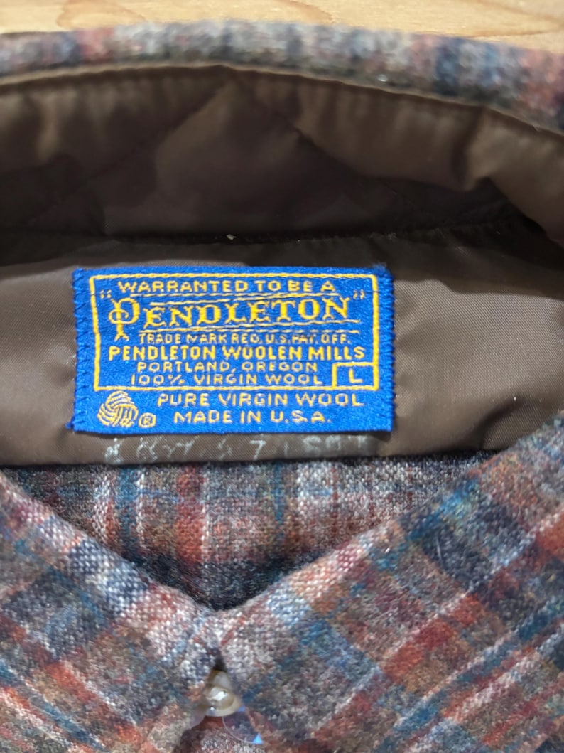 May include: Close-up of a Pendleton wool shirt label. The blue label reads "Warranted to be a Pendleton" and "100% Virgin Wool." The shirt has a plaid pattern in shades of brown, blue, and red. The label indicates the shirt is a size L and made in the USA.