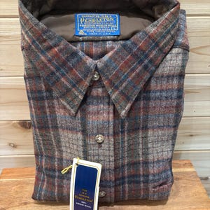 May include: A folded, button-down shirt with a plaid pattern in shades of brown, red, and blue. The shirt has a collar and a tag that reads "PENDLETON" and "100% VIRGIN WOOL". The shirt is made in the USA.