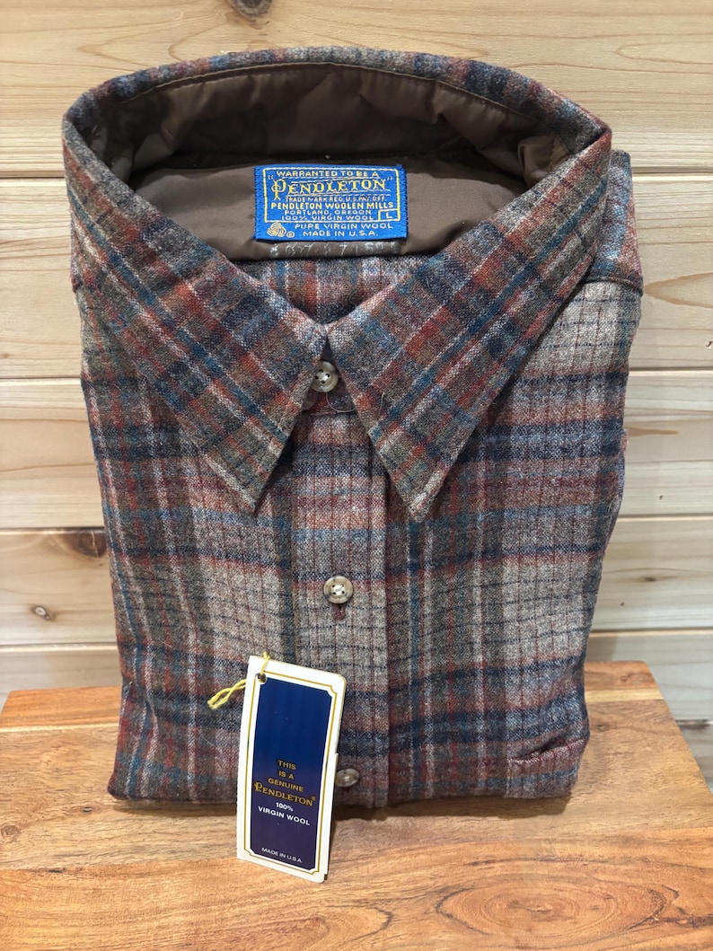 May include: A folded, button-down shirt with a plaid pattern in shades of brown, red, and blue. The shirt has a collar and a tag that reads "PENDLETON" and "100% VIRGIN WOOL". The shirt is made in the USA.