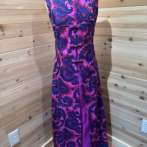 May include: A sleeveless floor-length dress featuring a vibrant paisley pattern in shades of magenta, royal blue, and red. The garment includes a high neckline and a central vertical panel adorned with three decorative fabric knot closures.