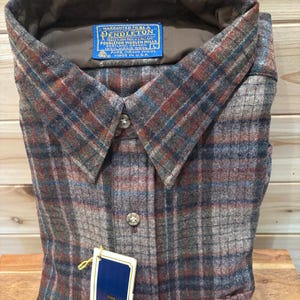 May include: A folded, button-down shirt with a plaid pattern in shades of brown, blue, and gray. The shirt has a collar and a blue tag that reads "PENDLETON" and "PURE VIRGIN WOOL". The shirt is made in the USA.