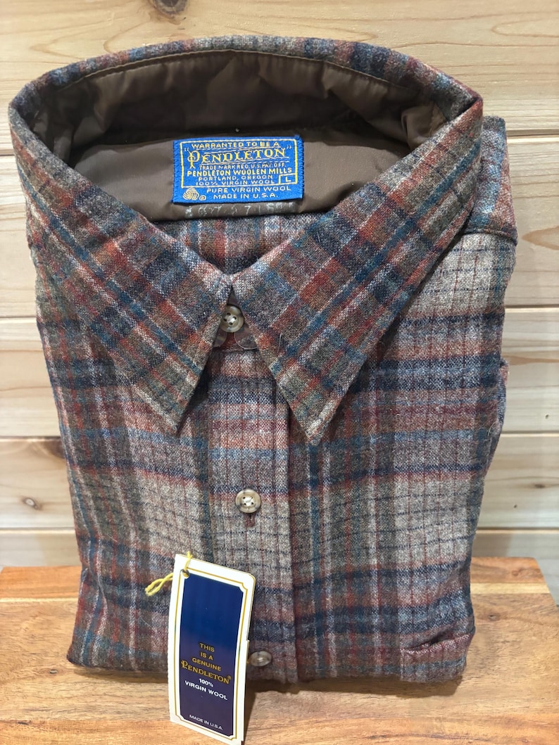 May include: A folded, button-down shirt with a plaid pattern in shades of brown, blue, and gray. The shirt has a collar and a blue tag that reads "PENDLETON" and "PURE VIRGIN WOOL". The shirt is made in the USA.