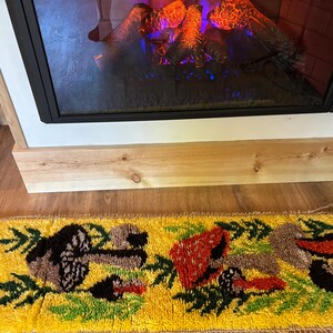 May include: A rectangular, yellow rug with a tufted design featuring birds and foliage. The rug is placed in front of a fireplace with a black frame and a wooden mantel. The rug's design includes red, brown, and green elements.