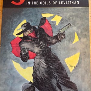 May include: Comic book cover for "The Shadow in the Coils of Leviathan." The artwork features a figure in a long black coat, hat, and red scarf, holding two guns. The title is in orange and black, with the author's names above.