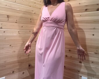 May include: A sleeveless, pale pink maxi dress with a V-neckline and a fitted waist. The dress has a flowing skirt and is made of a soft, lightweight fabric. The model is wearing the dress in front of a wood-paneled wall.