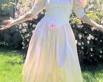 May include: A vintage-style, light pink dress with a white sequined bodice and a knee-length skirt. The dress features puff sleeves, a square neckline, and a pink flower accent at the waist. A bow adorns the shoulder.