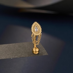 Puede incluir: Pendiente dorado con una parte superior en forma de lágrima, con una pequeña gema transparente y un borde decorativo. El pendiente tiene una cadena colgante y una pequeña bola en la parte inferior. La joya se muestra sobre un fondo oscuro.