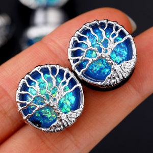 May include: Two round ear plugs with a tree of life design. The plugs have a silver-colored metal frame and a blue and green iridescent center. The tree design is in silver, and the plugs are set against a blurred background.