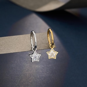 Puede incluir: Dos pendientes de aro, uno plateado y otro dorado, cada uno con un dije en forma de estrella. Los dijes de estrella son transparentes y facetados. Los pendientes se muestran sobre un fondo azul oscuro.