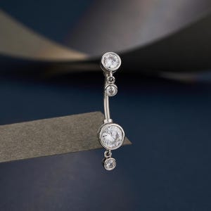 Puede incluir: Un piercing para el ombligo plateado con cuatro piedras preciosas redondas y transparentes. El diseño curvo de la barra presenta una gema grande en cada extremo y dos gemas más pequeñas en el centro. La joya está sobre un fondo azul oscuro.