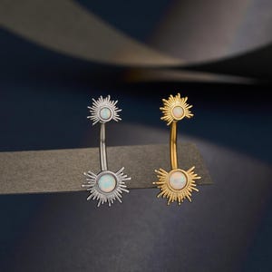 以下が含まれることがあります： 太陽のデザインのベリーボタンリングが2つ。1つはシルバーカラー、もう1つはゴールドカラーです。それぞれ、中心にオパールのような石と放射状のスパイクが付いています。ジュエリーは濃い青色の背景に表示されています。