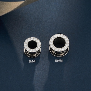 May include: Two silver ear gauges, each with a ring of clear crystals. One is marked "8mm", the other "10mm". The gauges are displayed on a dark blue surface. The 10mm gauge is approximately 0.4 inches in diameter.