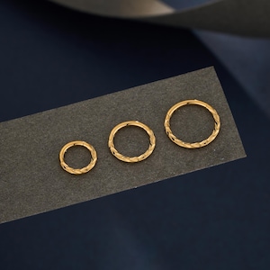 May include: Three gold-coloured hoop earrings of different sizes, each featuring a textured, woven-like design. The earrings are presented on a dark grey surface against a dark blue backdrop.