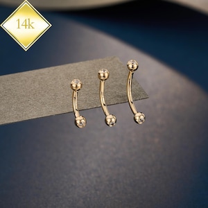 May include: Three gold-colored curved eyebrow rings with clear, round gemstones at each end. The jewelry is displayed on a gray surface against a dark blue background. A gold diamond-shaped tag with "14k" is in the upper left corner.