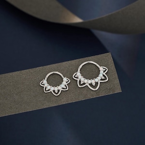 Può includere: Due orecchini a cerchio color argento con un design floreale. Ogni orecchino presenta un anello di piccole pietre bianche e un contorno a forma di fiore. Gli orecchini sono esposti su una superficie grigio scuro su sfondo blu scuro.