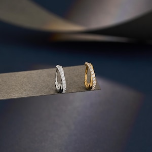 Puede incluir: Dos pendientes de aro, uno plateado y otro dorado, cada uno incrustado con pequeñas piedras preciosas transparentes. Los pendientes se muestran sobre una superficie gris sobre un fondo azul oscuro. El pendiente dorado está a la derecha y el plateado a la izquierda.