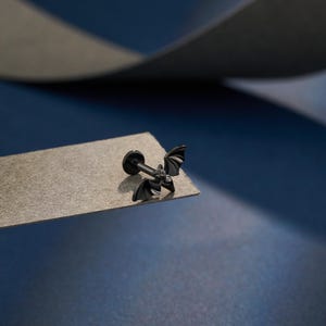 Op de afbeelding: Een zwarte lip piercing stud in de vorm van een vleermuis. De stud heeft een platte achterkant en een decoratief vleermuisontwerp met gedetailleerde vleugels. De sieraden zijn geplaatst op een grijze ondergrond met een blauwe achtergrond.