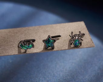 Pode incluir: Três brincos pretos com designs únicos: um morcego, uma estrela e uma aranha. Cada brinco apresenta uma incrustação verde escura e brilhante. Os brincos são exibidos em uma superfície de tom neutro contra um fundo azul escuro.