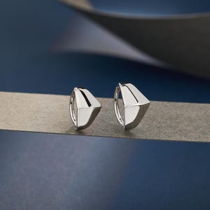 Puede incluir: Un par de pendientes de aro geométricos plateados. Cada pendiente presenta un diseño triangular con una superficie pulida y reflectante. Los pendientes se exhiben sobre una superficie gris sobre un fondo azul oscuro.