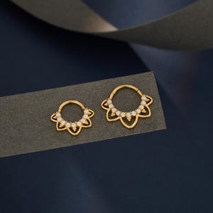 May include: Two gold-coloured, flower-shaped hoop earrings with small, white gemstones. The earrings are displayed on a dark grey surface against a dark blue background. The earrings are likely for ear or nose piercings.